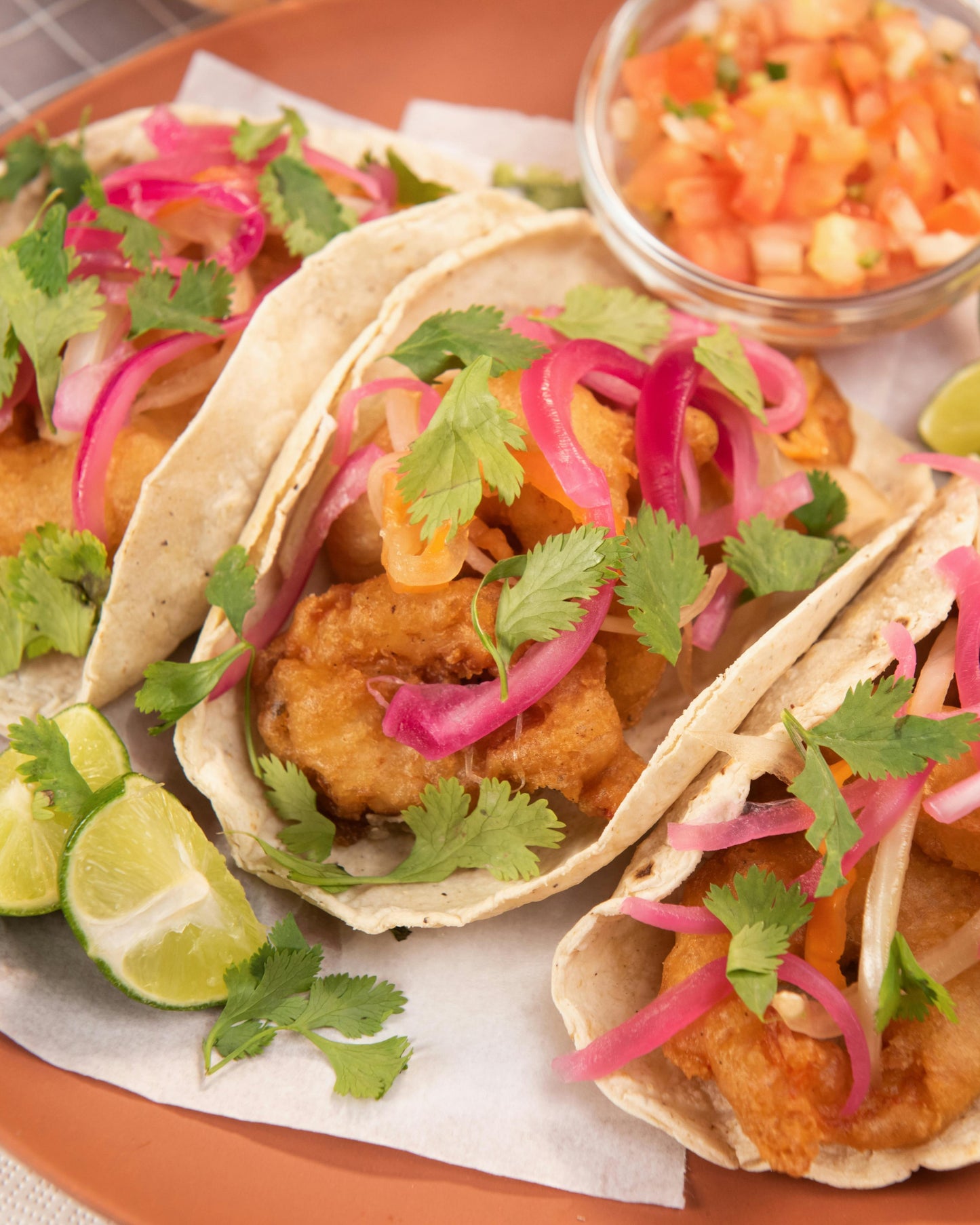 Orange plate with three fish tacos topped onion and cilantro with two slices of lime and a side of salsa